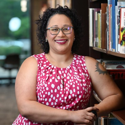 MLIS Alumna Mariela Siegert poses next to a bookshelf in a library. She has short, curly dark hair and is smiling. She is wearing glasses and a bright pink sleeveless top with a white polka dot pattern.