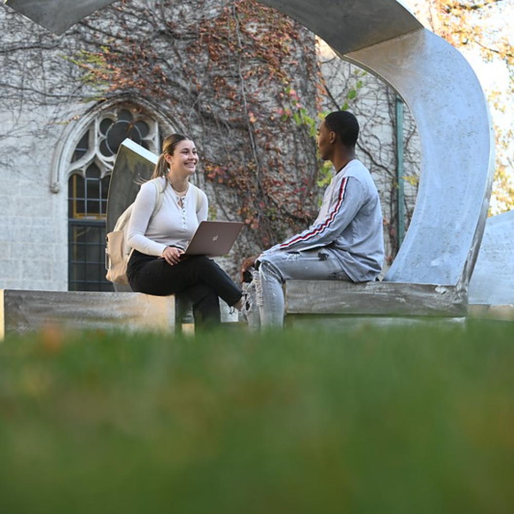 A white female student wearing a white, long-sleeved top and black pants, holds a laptop while seated on a metal sculpture on Dominican University's campus. She is smiling at a Black male student wearing a light gray shirt and gray pants, also seated on the same sculpture. 