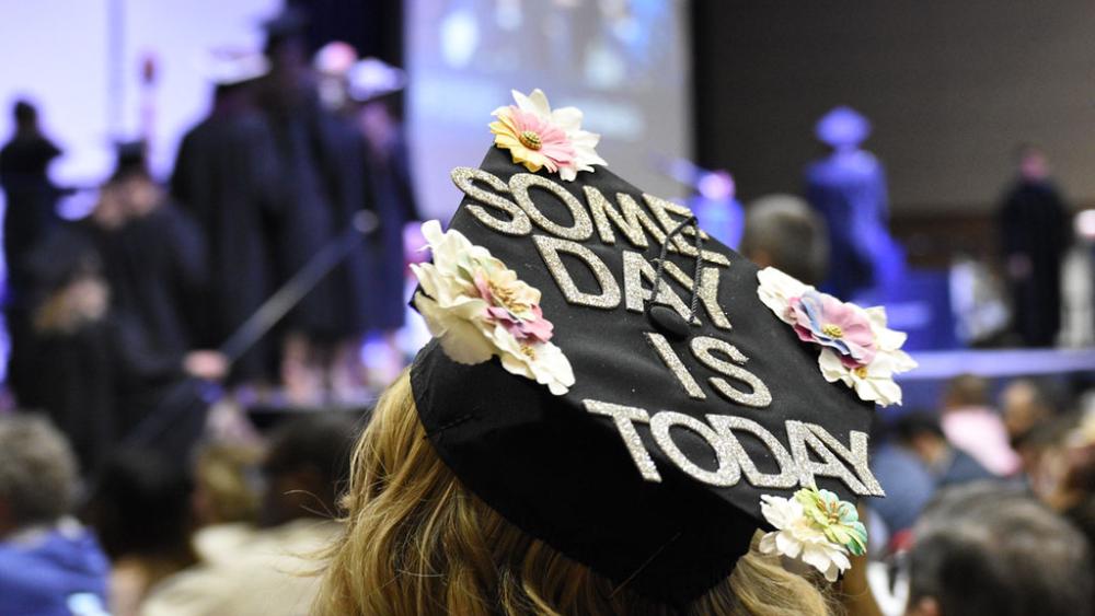 A student is seated at Dominican University's Commncement ceremony. The student's back is to the camera. The student's graduation cap is decorated with flowers and the words Some Day is Today.