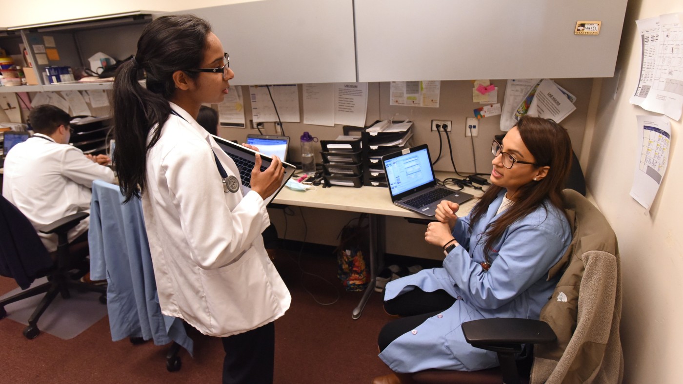 Two female students are speaking to each other in an office setting. One is standing, wearing a white lab coat and a stethoscope around her neck. The other student is seated in front of a computer, wearing a light blue lab coat. A male student sits to their left, looking at a laptop.
