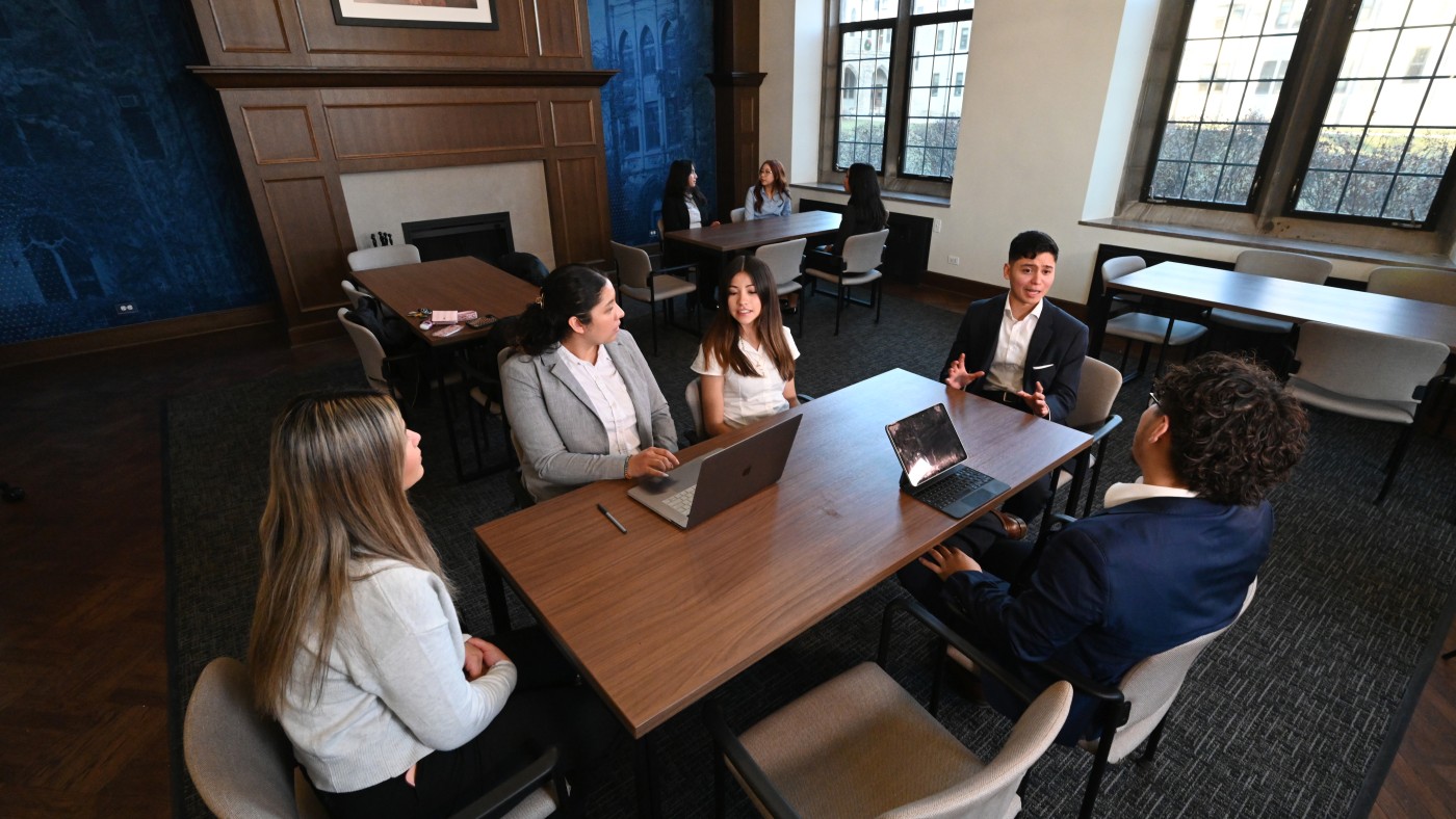  Students in business attire sit around a table in an academic study room, discussing with laptops open. Other small groups talk in the background.