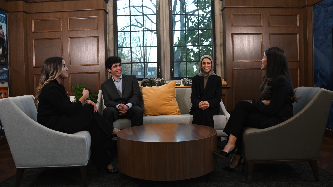 Students sit in armchairs around a low table in a wood-paneled room on campus.
