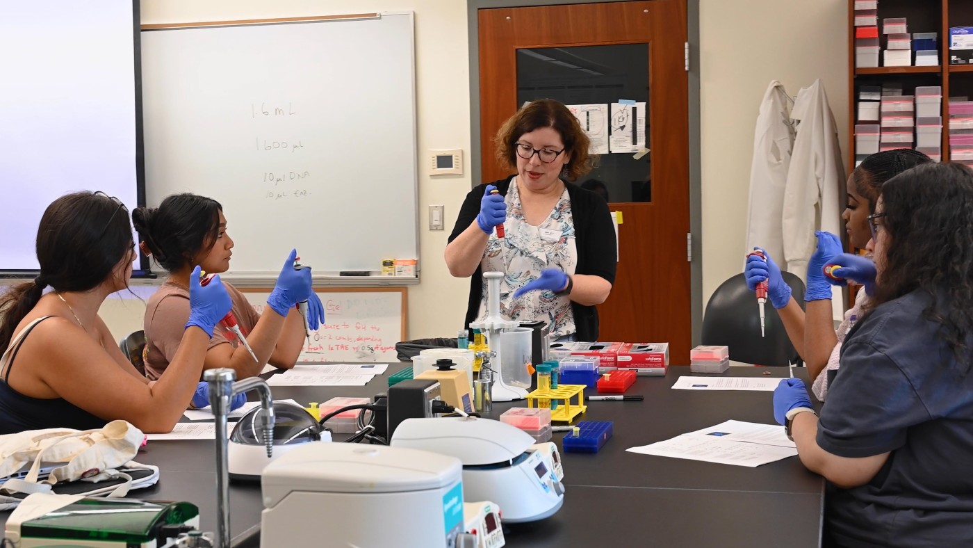 Students seated around a lab table watch their professor demonstrate a lab process.