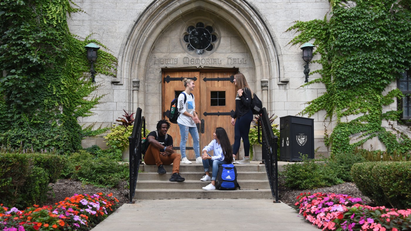 A group of four students talking in front of the Lewis Hall exterior door. Two students stand in front of the door. Two students are seated on the steps leading up to the door.