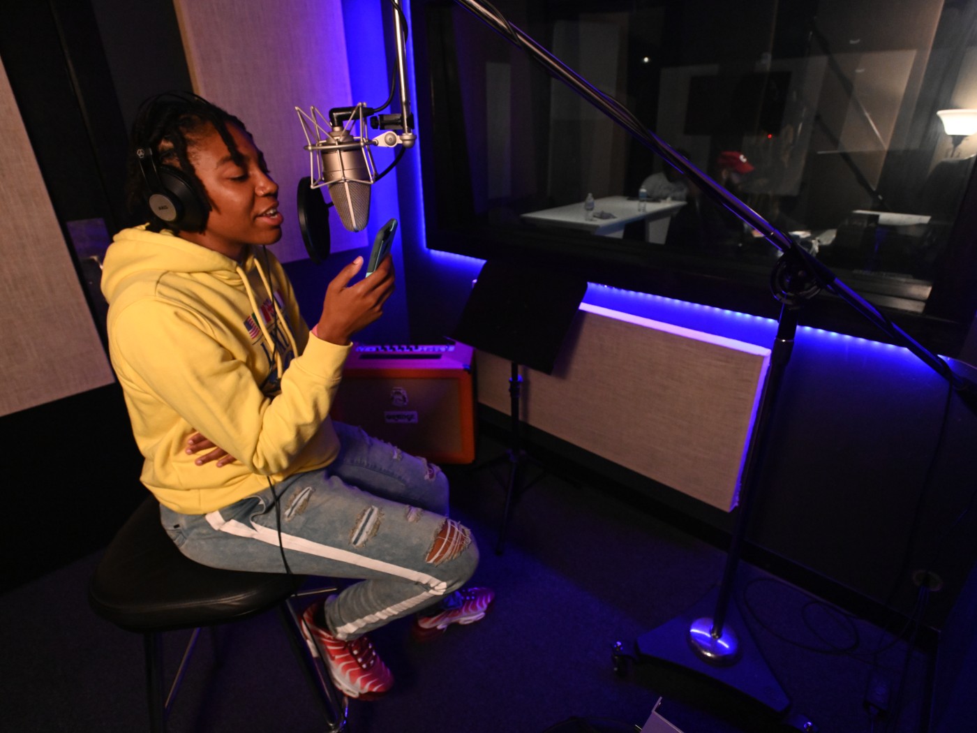 Dominican student Timiya Ray is wearing a yellow hoodie and ripped jeans sits on a stool in a dimly lit recording studio, singing into a large microphone. Timiya wears headphones and holds a phone, reading lyrics. The room is lit with blue LED lights and features soundproofing panels, a window to the control room, and recording equipment.