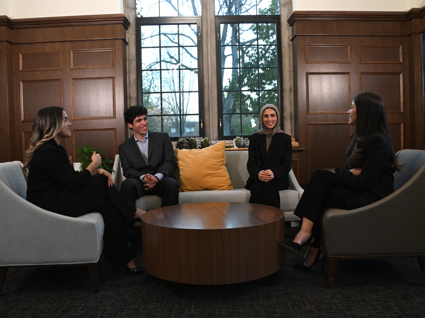 Students sit in armchairs around a low table in a wood-paneled room on campus.