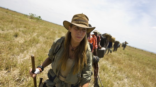 Mireya Mayor - A womon in bush gear and a hat hiking through the Savanna.